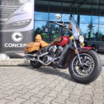 Czerwony motocykl Indian Scout z brązowymi sakwami skórzanymi, zaparkowany przed budynkiem.