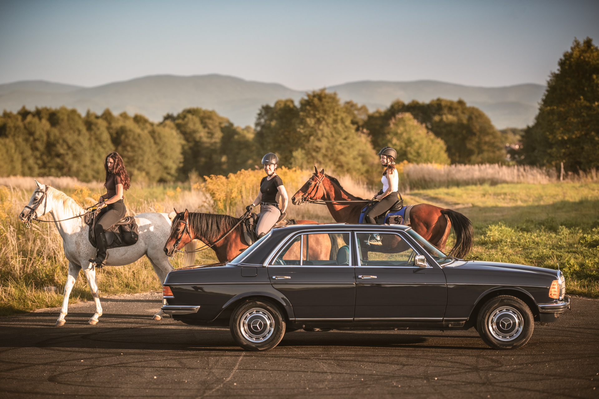 Klasyczny czarny Mercedes-Benz W123 na tle natury z kobietami na koniach.