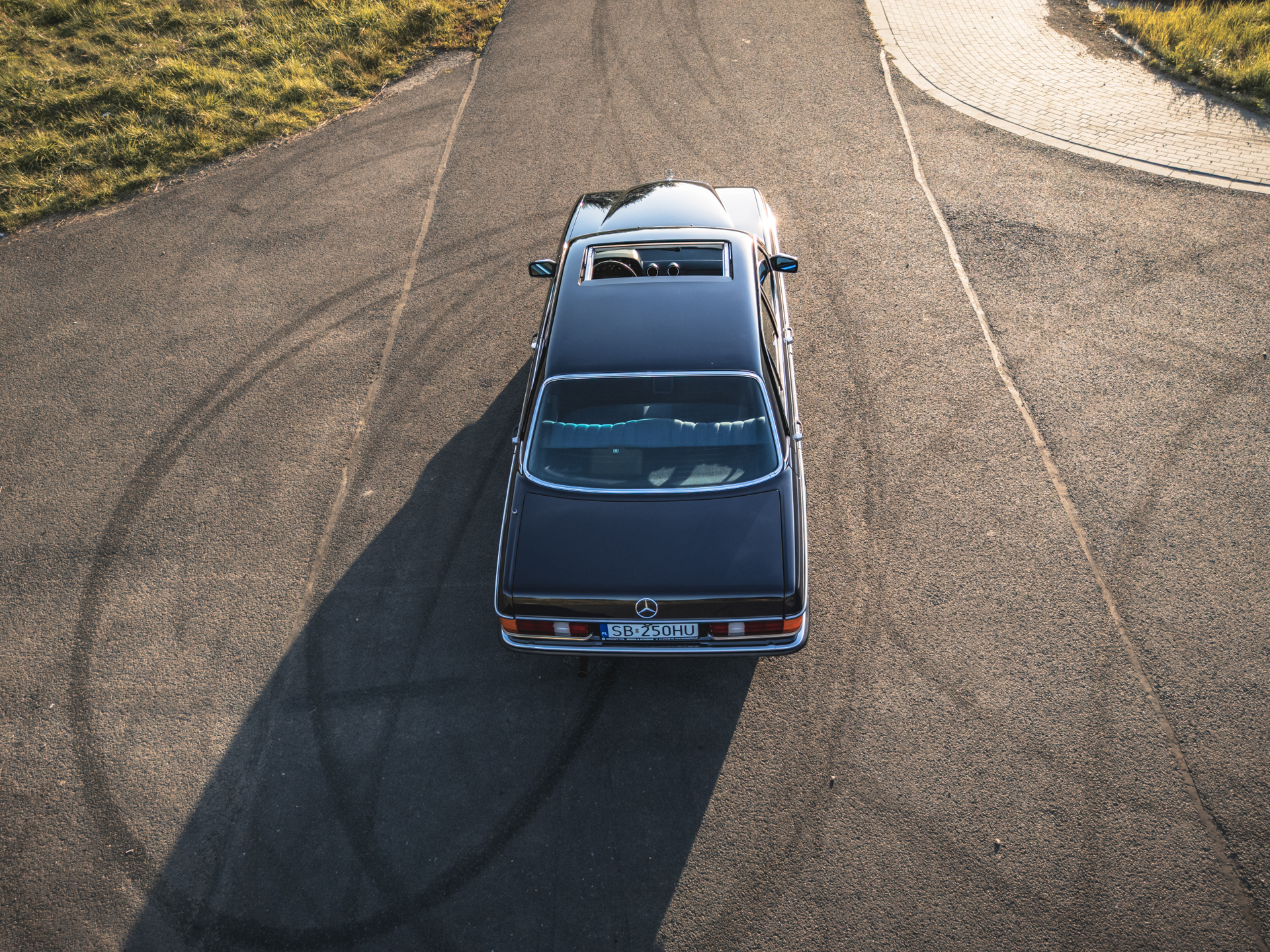 Czarny Mercedes-Benz W123 z szyberdachem widziany z góry na drodze.