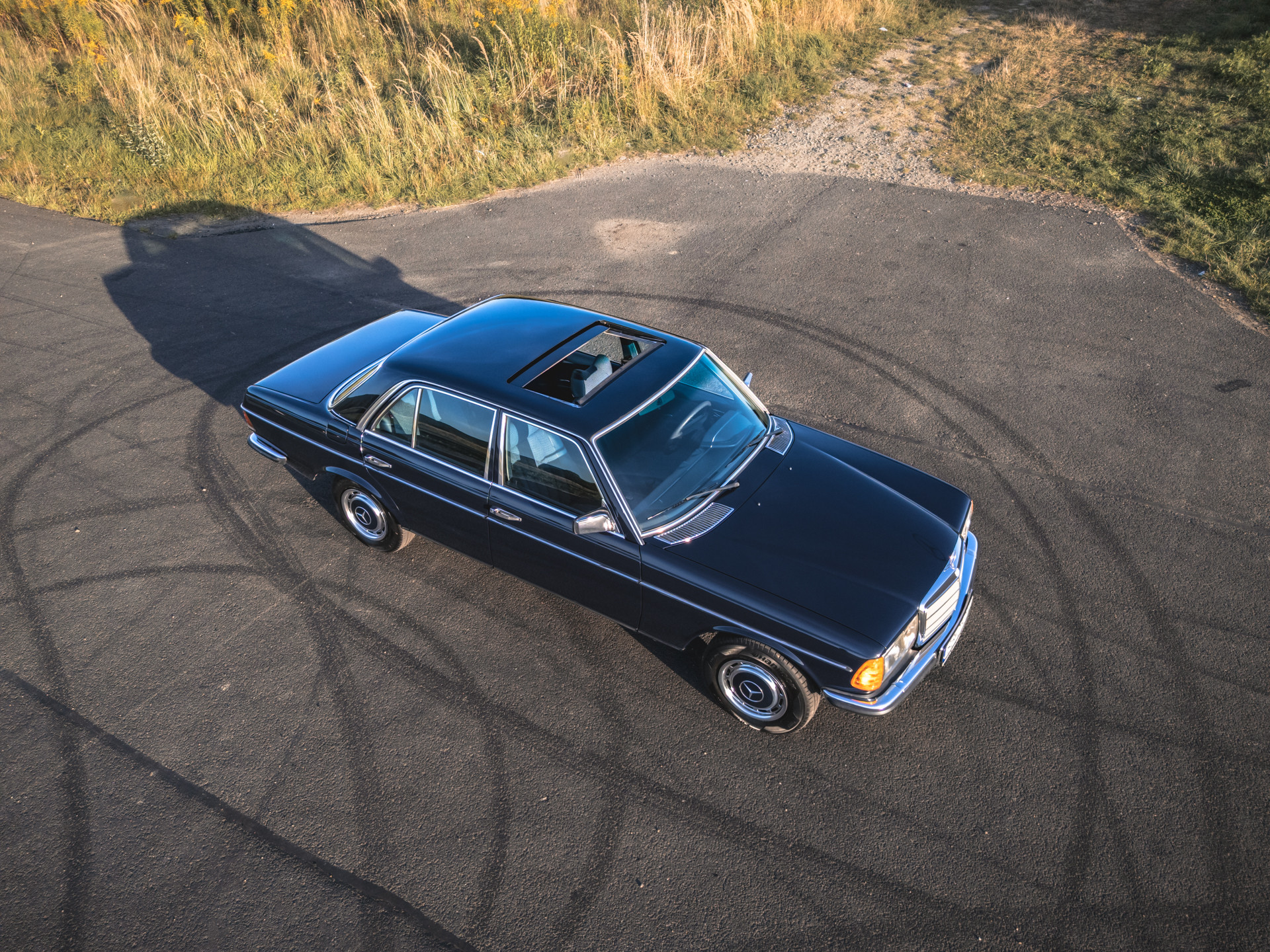 Ciemnoniebieski sedan Mercedes-Benz W123 z szyberdachem na asfaltowej drodze.