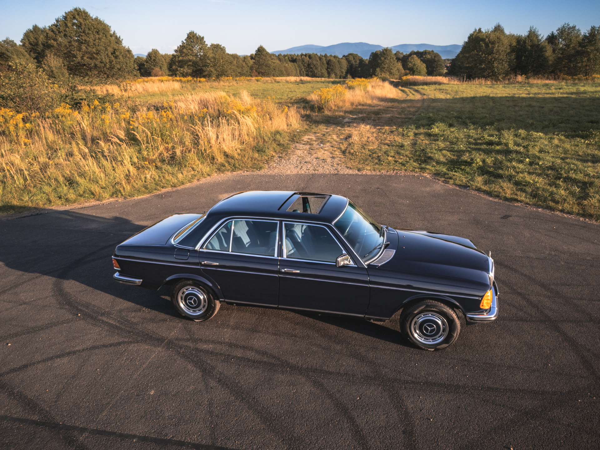 Ciemnoniebieski Mercedes-Benz W123 sedan z szyberdachem na tle krajobrazu.